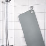 Gray bath mat hangs on wall hook in tiled shower, secured by suction cups.