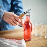 Person opens a bottle with a dual bottle/can opener, its wooden handle providing a firm grip.