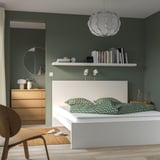 Modern bedroom with olive green walls, white bed, green pillows, wooden dresser, round mirror, bookshelf, hanging light, and chair.