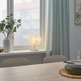 A modern dining area with a small white VINDKAST table lamp, plates, and chairs by a window. Light and airy decor.