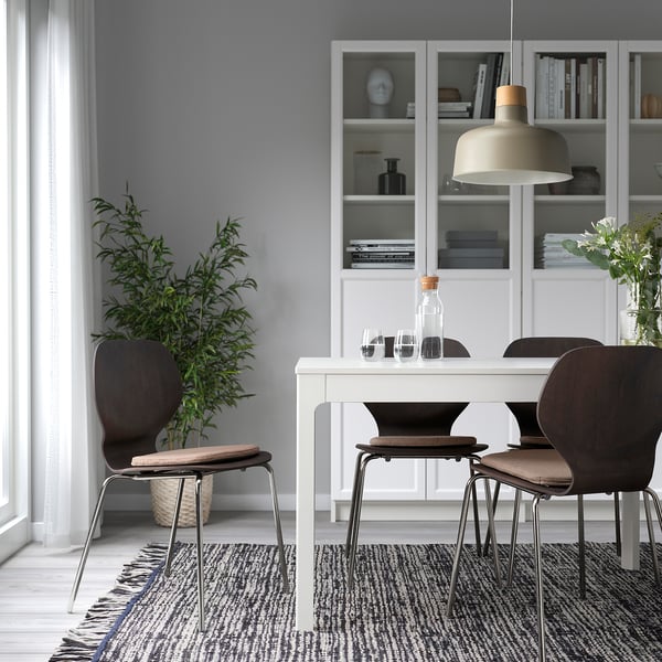 Configuration de salle à manger moderne avec des coussins de chaise ÄLVGRÄSMAL en beige, positionnés sur des chaises marron foncé autour d’une table blanche sur un tapis à motifs sous une lampe suspendue.