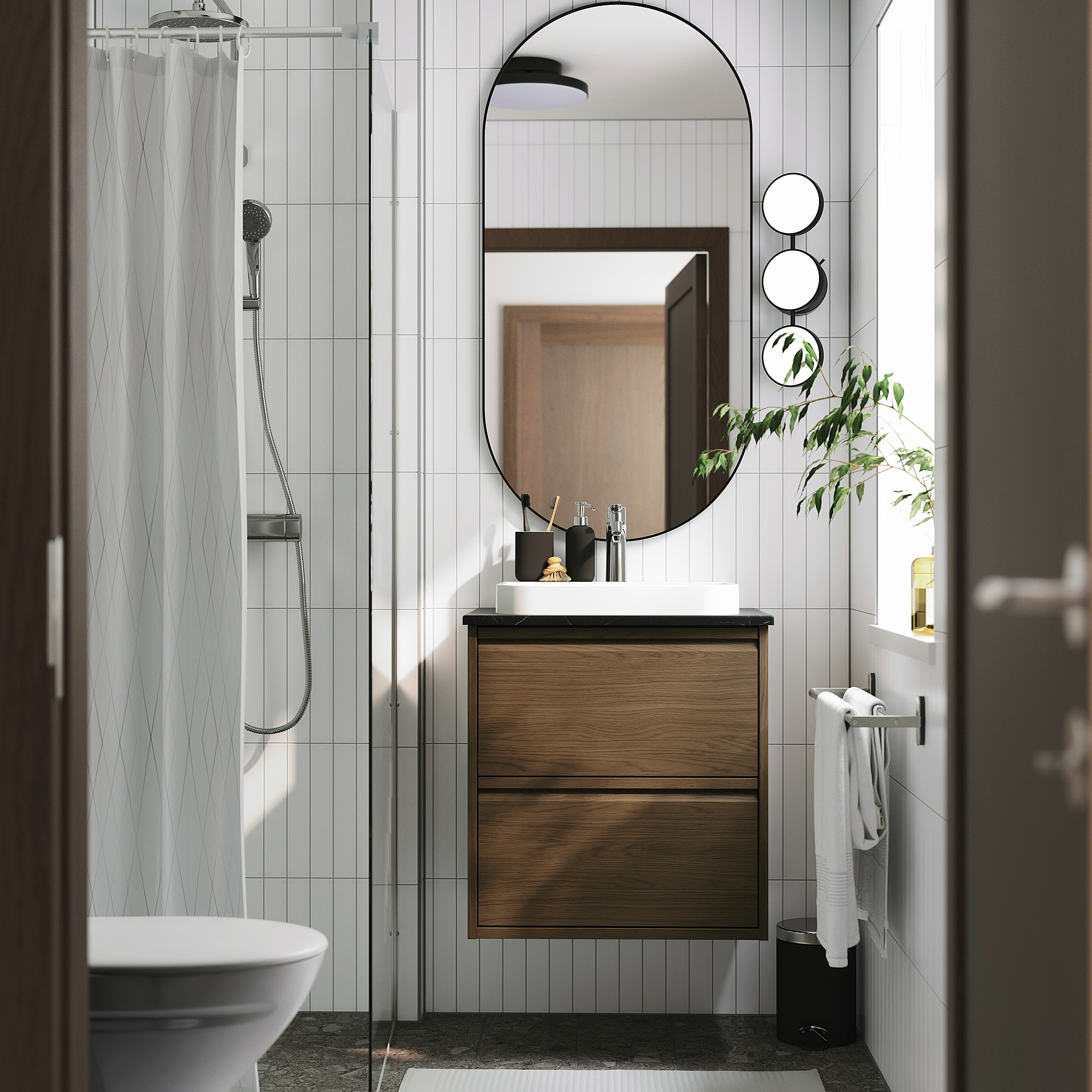 Salle de bain moderne avec meuble-lavabo en bois ÄNGSJÖN, lavabos blancs, douche en verre et miroir rond. Les plantes ajoutent du vert.