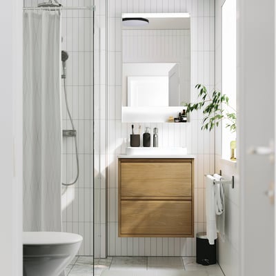 Salle de bain moderne avec armoire en bois, vanité, lavabo, douche, miroir et plante.