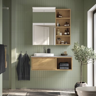 Salle de bain moderne et verte avec armoire en bois, lavabo blanc, miroir, serviettes et plante.