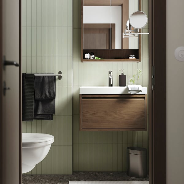 Salle de bain moderne avec carrelage vert clair, lavabo blanc, armoire en bois sombre, serviettes noires et accents métalliques. La vanité s’adapte parfaitement sous un grand miroir.