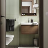 Salle de bain moderne avec carrelage vert clair, lavabo blanc, armoire en bois sombre, serviettes noires et accents métalliques. La vanité s’adapte parfaitement sous un grand miroir.