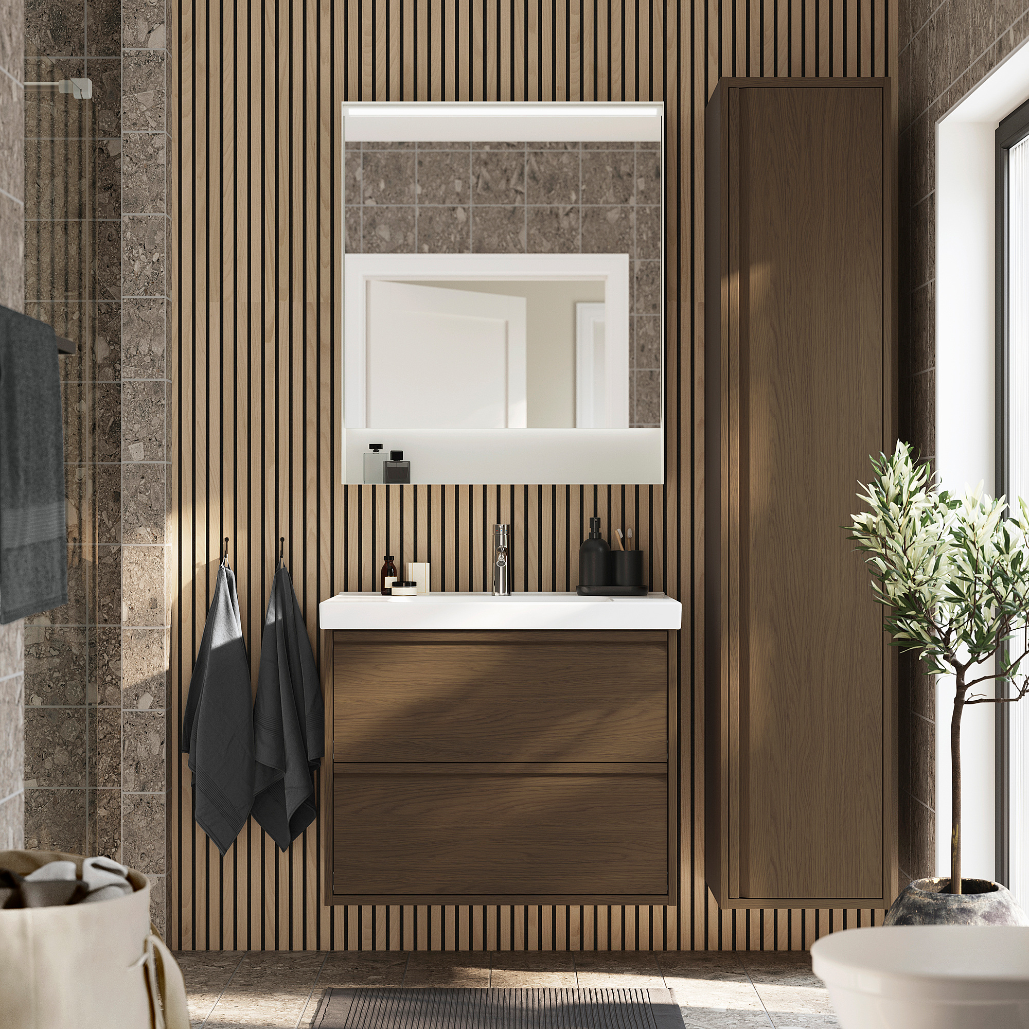 Salle de bain moderne avec armoire en noyer brun ÄNGSJÖN, grand miroir, lavabo blanc, serviettes grises et plante verte.