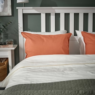 Taie d’oreiller orange rouille ÄNGSLILJA, coton doux, convient à un oreiller standard, sur un lit douillet, avec des draps blancs, un panier et une table de chevet.