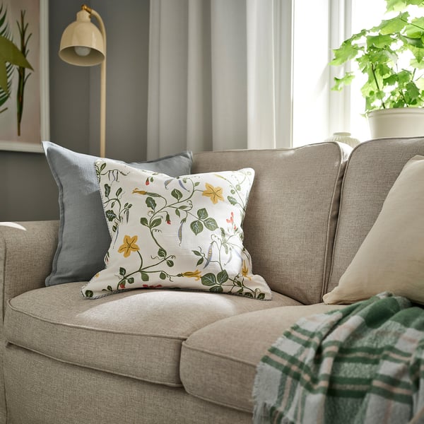 Canapé beige, coussin floral, oreiller gris, couverture verte, lampe, plante.