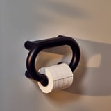 Porte-papier toilette noir au design arrondi, monté sur un mur gris. Contient un rouleau de papier toilette blanc.