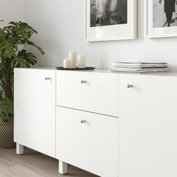 Armoire blanche moderne avec des boutons en métal, contenant des plantes, des bougies et des livres, positionnée sous des œuvres d’art murales encadrées.