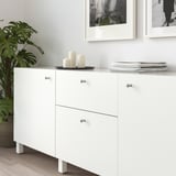 Armoire blanche moderne avec des boutons en métal, contenant des plantes, des bougies et des livres, positionnée sous des œuvres d’art murales encadrées.