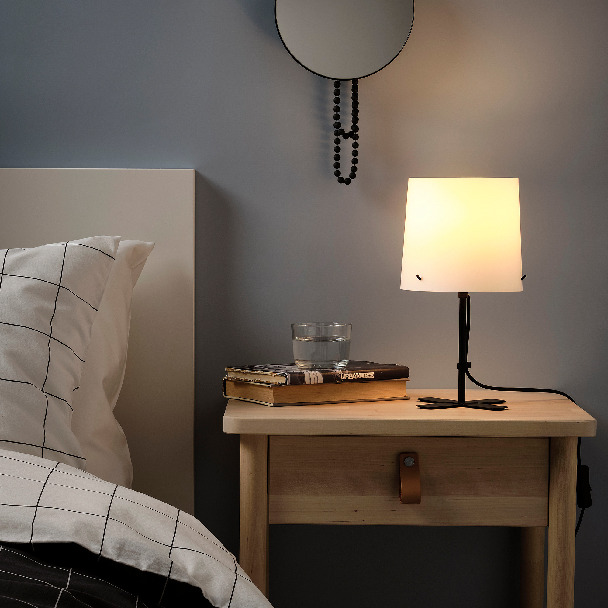Chevet avec lampe BALAST, livres, verre, table de chevet, lit, coussin grillagé, mur gris, ornement suspendu.
