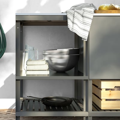 Unité de cuisine extérieure ouverte avec pieds réglables. Plan de travail en acier inoxydable. Étagères avec serviettes empilées, pichet et grand bol. Poêle sur l’étagère ci-dessous.