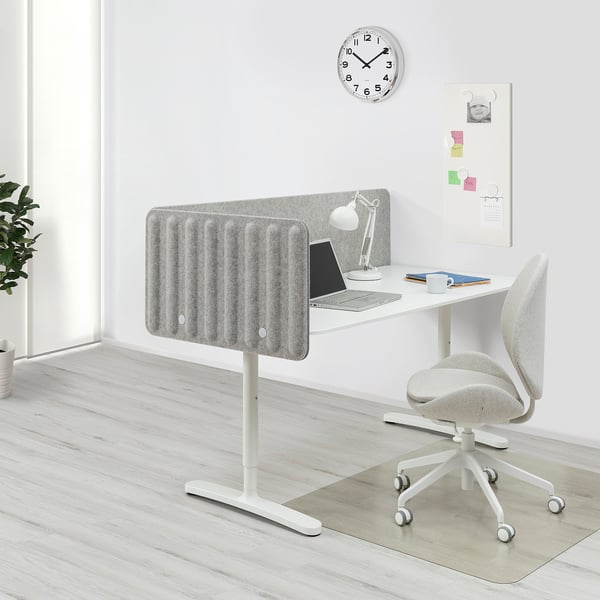 Bureau à domicile moderne avec bureau BEKANT, chaise grise, gestion des câbles et écran de confidentialité. Horloge et tableau d’affichage sur mur.