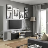 Salon moderne avec banc TV BESTÅ en noir et blanc. Télévision montée sur mur avec des cadres d’art. Lampadaire, table basse et rideaux gris.