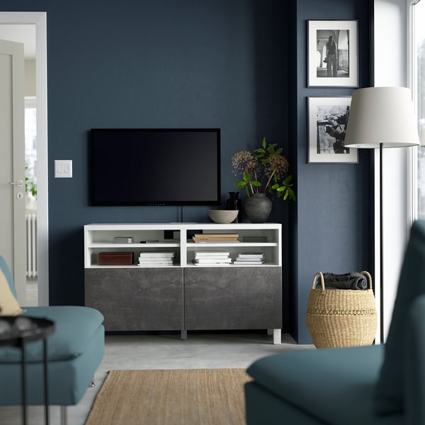 Salon moderne avec banc TV BESTÅ, télévision à écran plat, canapé sarcelle, panier, murs bleu foncé, photos, grande lampe, livres, bol, fleurs.