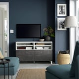 Salon moderne avec banc TV BESTÅ, télévision à écran plat, canapé sarcelle, panier, murs bleu foncé, photos, grande lampe, livres, bol, fleurs.