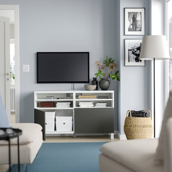 Salon moderne avec meuble TV BESTÅ blanc, télévision à écran plat, canapé blanc, tapis bleu, panier en osier et photos encadrées.