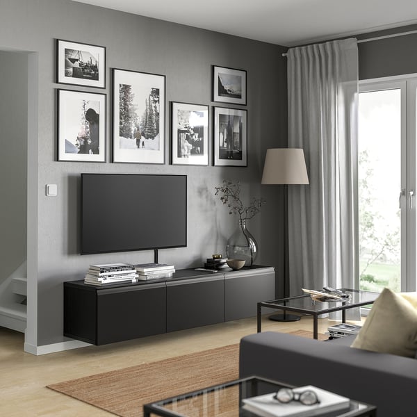 Salon moderne avec banc TV BESTÅ noir, télévision grand écran, photos encadrées, table basse en verre, canapé gris et grande fenêtre avec rideaux.