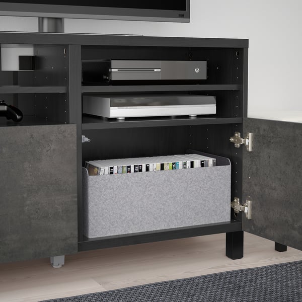 Banc TV BESTÅ noir avec porte ouverte révélant une boîte grise contenant des boîtiers de jeux et des consoles. Charnières à fermeture douce visibles.
