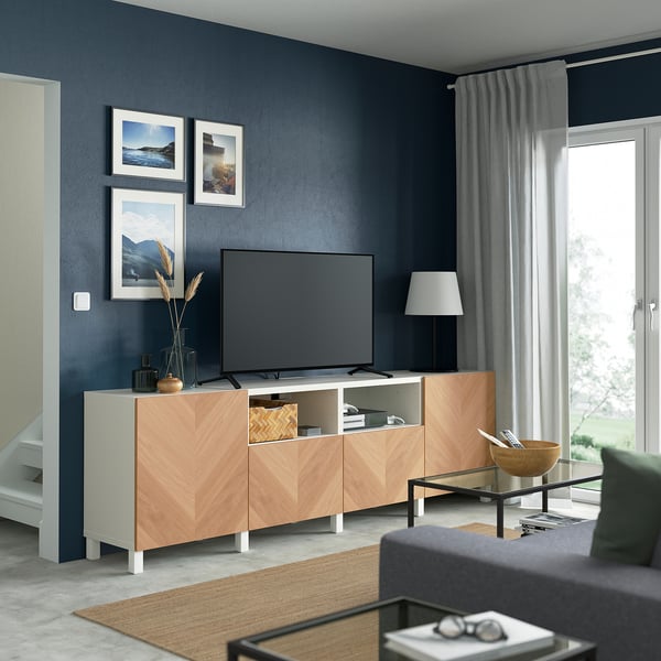 Salon moderne avec banc TV BESTÅ en bois clair, mur sombre, canapé gris, grande télévision à écran plat et objets de décoration.
