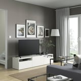 Salon moderne avec meuble tv BESTÅ, tv, décoration et plantes. Table basse en verre, canapé gris et grande fenêtre avec rideaux.