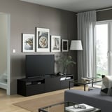 Salon moderne avec banc de télévision, lampe et tapis BESTÅ. Rideaux gris, art, table basse avec des livres et télécommande, canapé confortable.