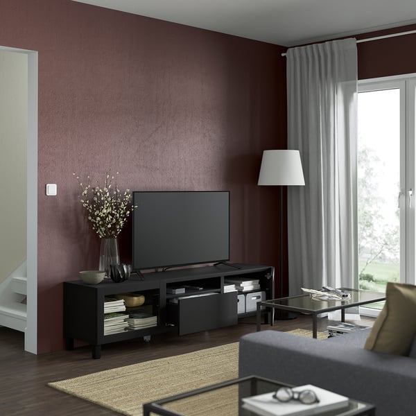 Salon moderne aux murs rouge foncé, banc TV BESTÅ, télévision à écran plat, fleurs séchées, lampe, table basse en verre, canapé.