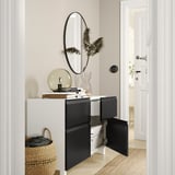 Entrée moderne avec buffet BESTÅ blanc et noir, portes ouvertes révélant des rangements, surmonté d’un décor, d’un miroir rond et d’un panier tressé.