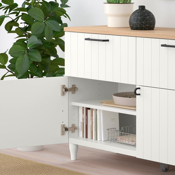 Unité de rangement moderne avec étagère ouverte contenant des livres et un panier, une armoire fermée au-dessus et des objets décoratifs sur le dessus. Des plantes et un bol sont à proximité.
