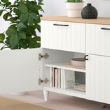 Unité de rangement moderne avec étagère ouverte contenant des livres et un panier, une armoire fermée au-dessus et des objets décoratifs sur le dessus. Des plantes et un bol sont à proximité.