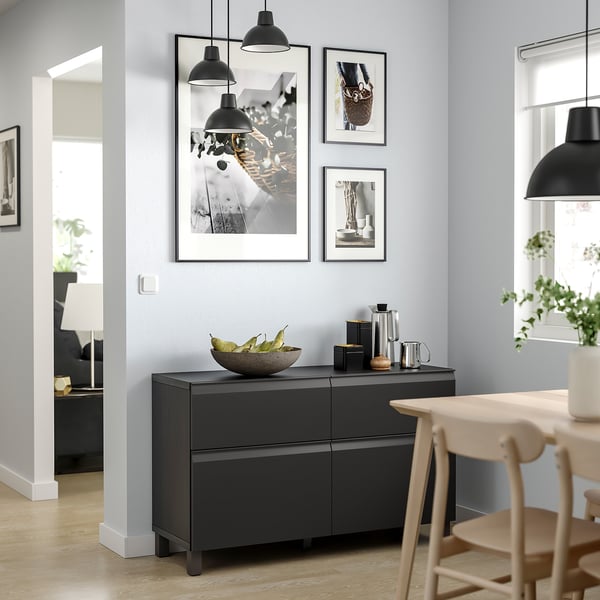Salle à manger moderne avec buffet BESTÅ noir, suspensions et chaises en bois. Le mur présente des œuvres d’art encadrées.
