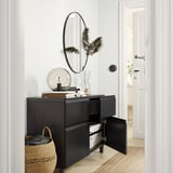 Buffet BESTÅ noir avec porte ouverte et tiroir, avec miroir, lampe globe, livres, vase avec plante, panier avec couverture dans une pièce moderne.