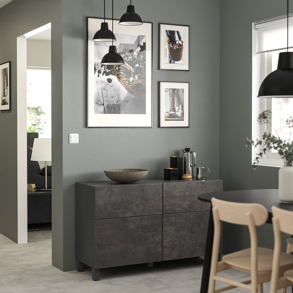 Salon moderne avec unité de rangement BESTÅ grise, chaises en bois et lampes suspendues. Œuvre d’art sur mur végétal.