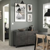 Salon moderne avec unité de rangement BESTÅ grise, chaises en bois et lampes suspendues. Œuvre d’art sur mur végétal.