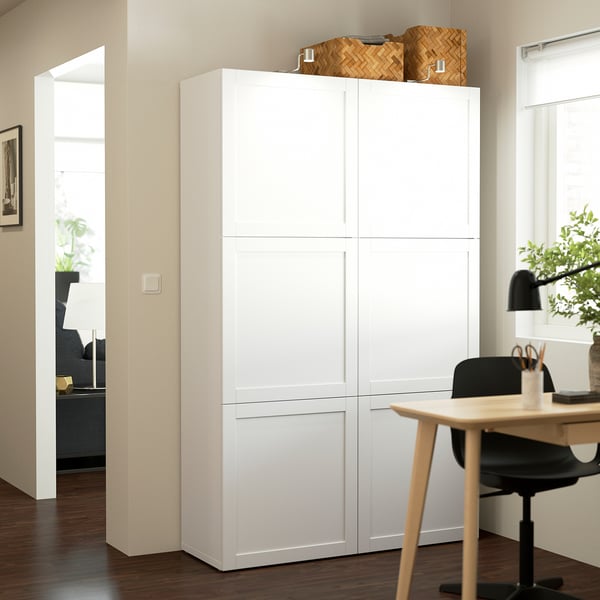 Unité de rangement BESTÅ blanche dans l’espace de vie, plus haute que le bureau adjacent, avec des paniers tressés sur le dessus.