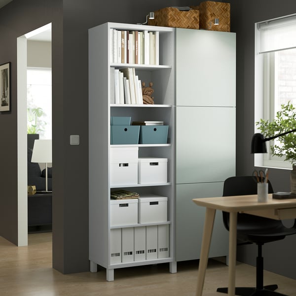 Unité de rangement BESTÅ blanche avec étagères réglables, boîtes de rangement et livres, plus haute que les meubles à proximité.