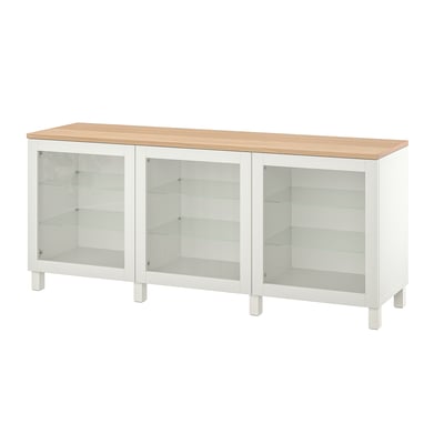 Un buffet IKEA BESTÅ blanc avec trois portes vitrées et un plateau en bois. L’armoire a un design moderne avec des étagères réglables pour le rangement.