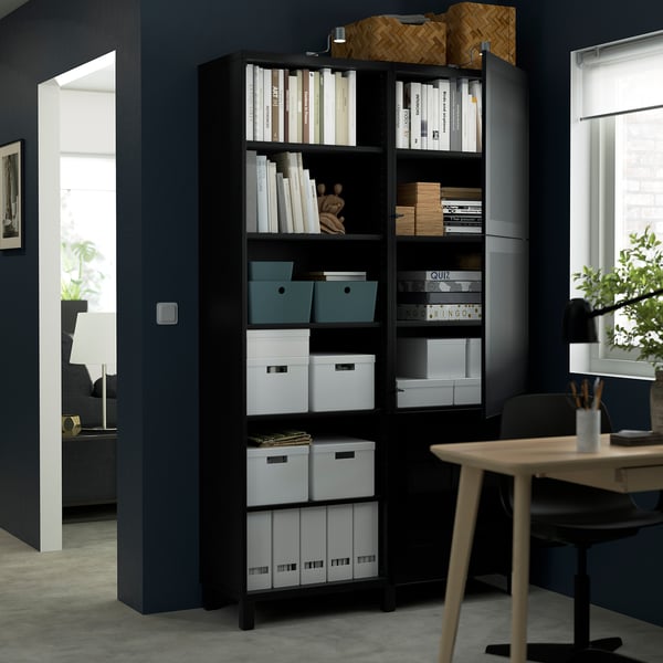 Unité de rangement BESTÅ noire avec étagères et armoires, contenant des livres, des boîtes et des objets décoratifs. Personnalisable et fixé au mur pour plus de sécurité.