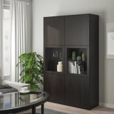 Unité de rangement BESTÅ noire avec porte vitrée exposant des vases et des livres. Sa grande taille s’intègre parfaitement dans le salon, à côté d’une table près d’une fenêtre.