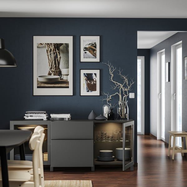 Salle à manger moderne avec unité de rangement BESTÅ gris foncé, chaises blanches et œuvres d’art encadrées sur un mur bleu foncé.