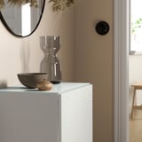 Le plateau en verre blanc BESTÅ se trouve entre un vase et un bol, se reflétant dans un miroir rond. Élégant et moderne, il complète la porte en bois adjacente et la table du fond.