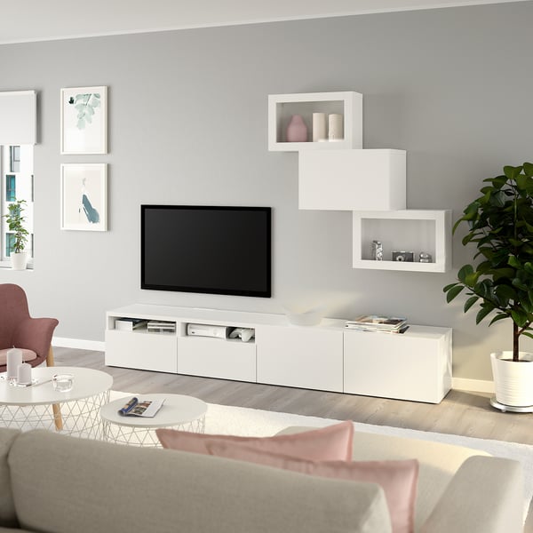 Salon moderne avec rangement TV BESTÅ blanc, télévision à écran plat, canapé beige, oreillers roses, table basse ronde, plante verte et art minimaliste.