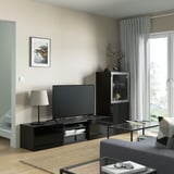 Salon moderne avec rangement TV noir BESTÅ, télévision à écran plat, armoire latérale noire, table en verre, rideaux gris et parquet.