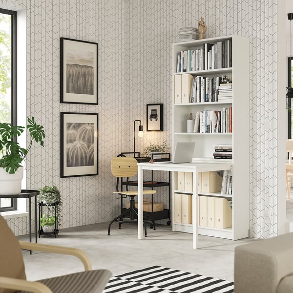 Bureau à domicile moderne Bibliothèque BILLY avec bureau, blanc. Des cadres muraux, un ordinateur portable et des plantes rehaussent l’espace bien rangé et élégant.