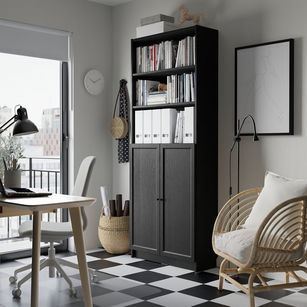 Bureau à domicile avec bibliothèque BILLY, noir, rempli de livres et de décoration. Horloge blanche, sol en damier, chaises noires.