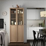 Bibliothèque avec portes vitrées dans la cuisine moderne. Cheval en bois au-dessus, évier blanc, chaises noires, horloge murale, lampe suspendue.