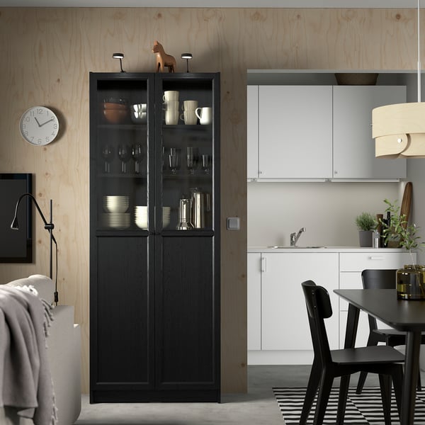 Armoire BILLY noire dans la cuisine minimaliste, près de l’ensemble de salle à manger blanc, avec des portes vitrées montrant la vaisselle.