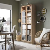Bibliothèque BILLY en bois clair avec portes vitrées, près d’une chaise blanche et d’une petite table, décorée d’assiettes et de vases.